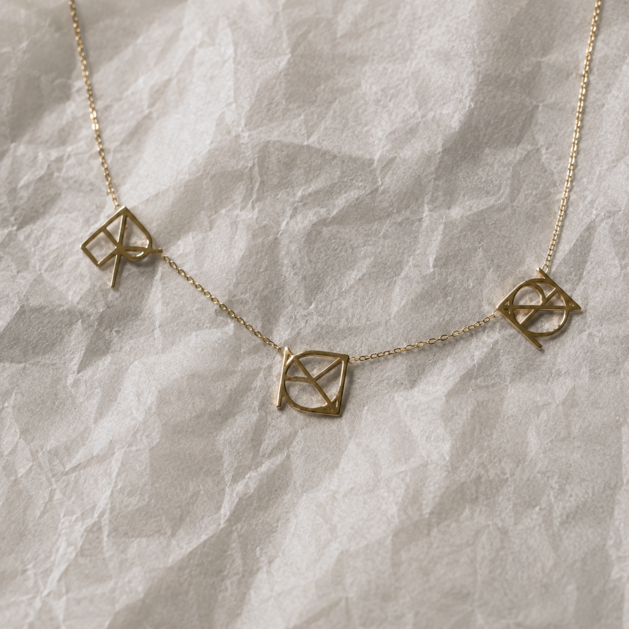 A top-down studio product shot of the Danibydsgn three symbol pendant necklace in 9k solid gold. This made to order modern heirloom necklace features three personalized uppercase geometric charms handcrafted from a customer's meaningful names, words, or dates, beautifully laid out across a textured white crinkled paper background.