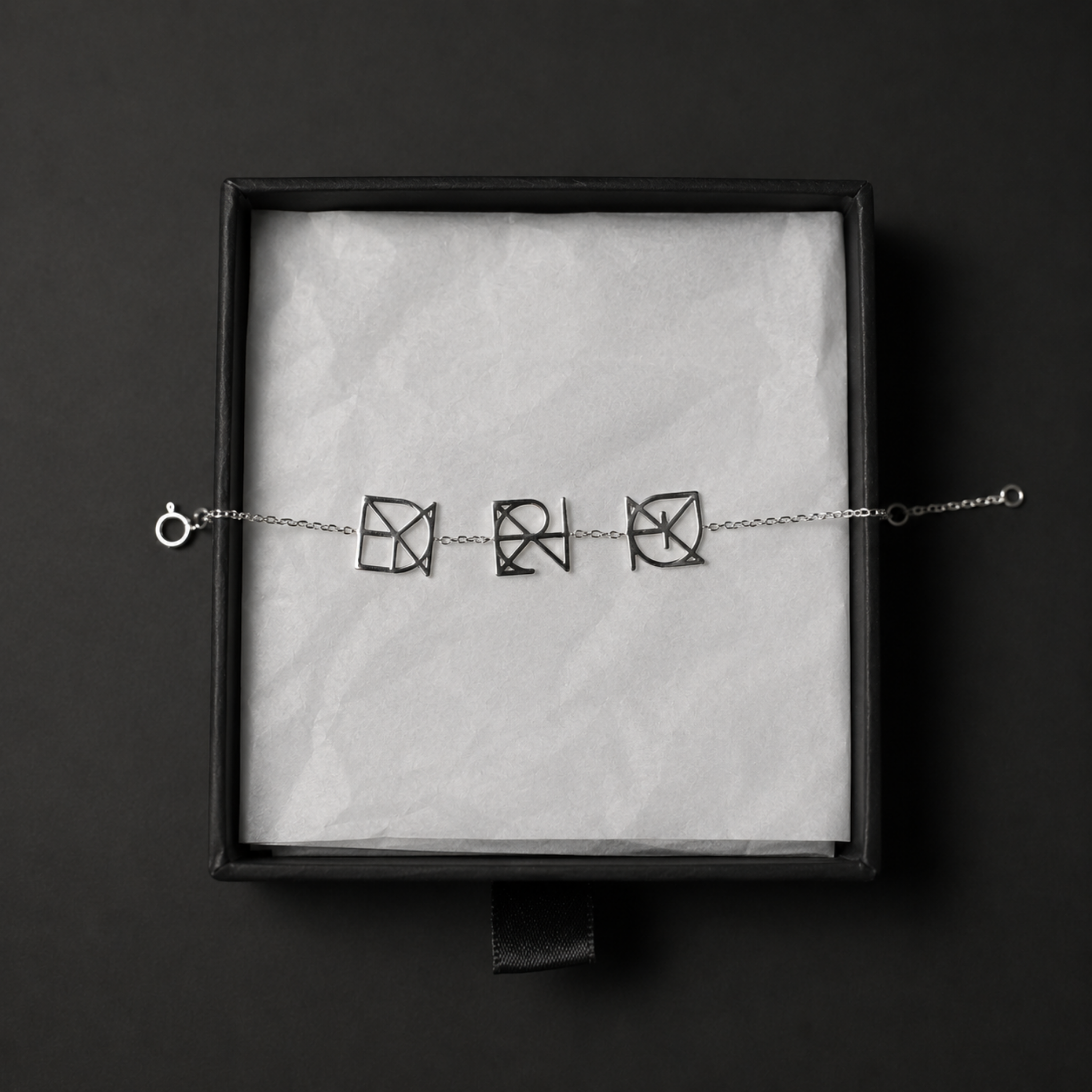 A top-down product shot of a Danibydsgn three symbol pendant bracelet in 925 sterling silver laid flat inside a black jewelry gift box. This personalized meaningful jewelry features three cut-out, uppercase geometric charms made exclusively from a custom fineline Danibydsgn design, beautifully arranged on textured white tissue paper.
