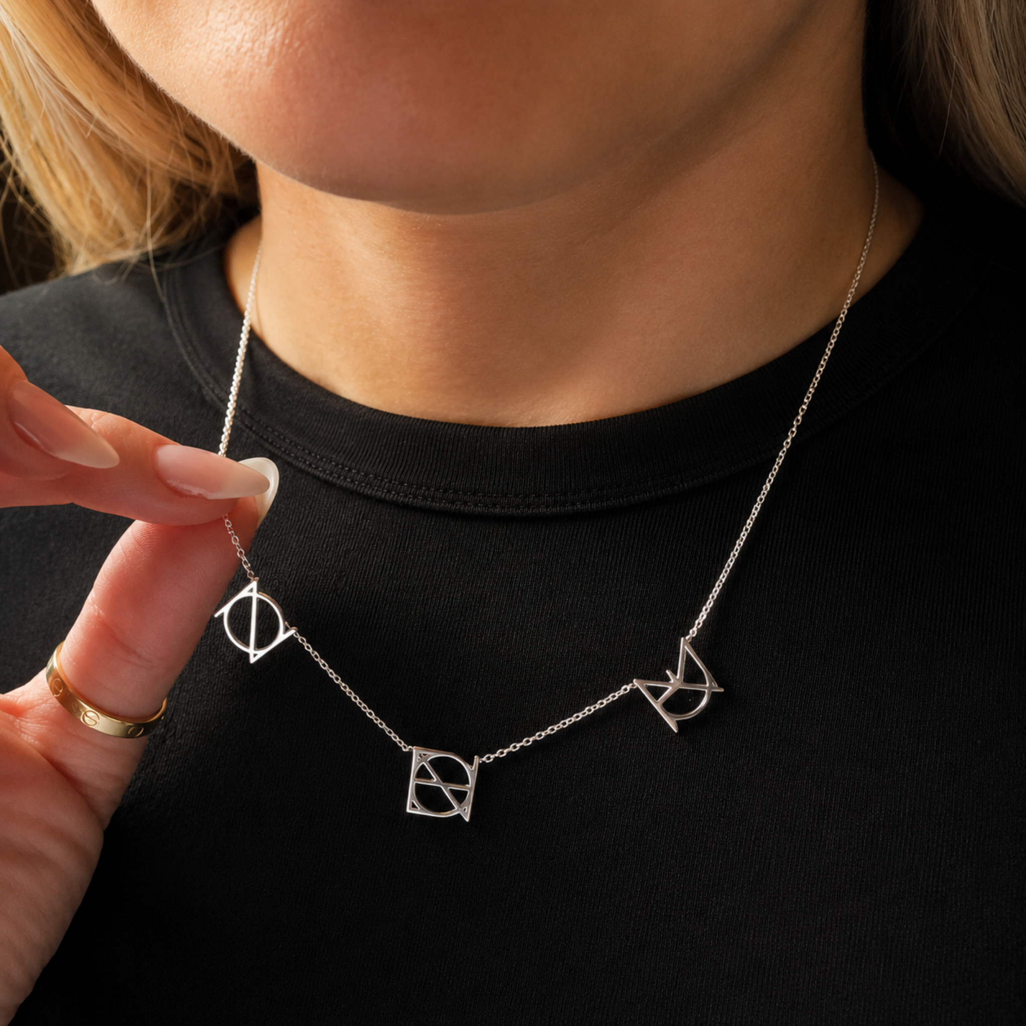 A close-up lifestyle image of a model wearing the Danibydsgn three symbol pendant necklace in 925 sterling silver against a black t-shirt. The model's fingers gently lift the side of the delicate silver chain to highlight three personalized uppercase geometric charms handcrafted from a custom fine-line design.