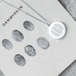 The Danibydsgn Three Fingerprint Coin Necklace in 925 Sterling Silver placed on a fingerprint design sheet — a symbolic connection between loved ones captured in precious metal, representing three hearts united through touch.