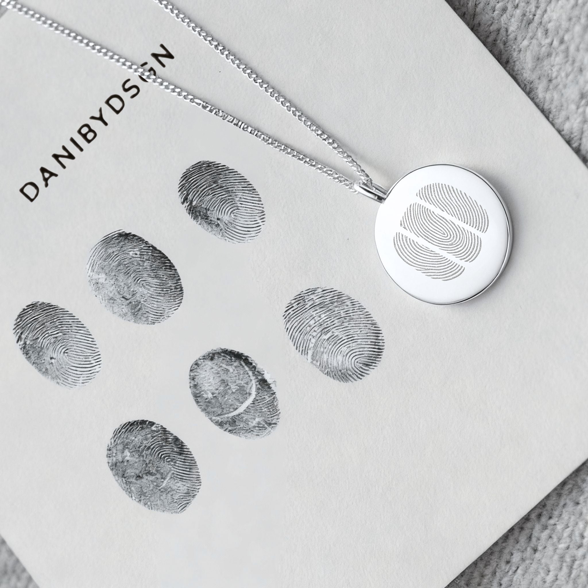 The Danibydsgn Three Fingerprint Coin Necklace in 925 Sterling Silver placed on a fingerprint design sheet — a symbolic connection between loved ones captured in precious metal, representing three hearts united through touch.