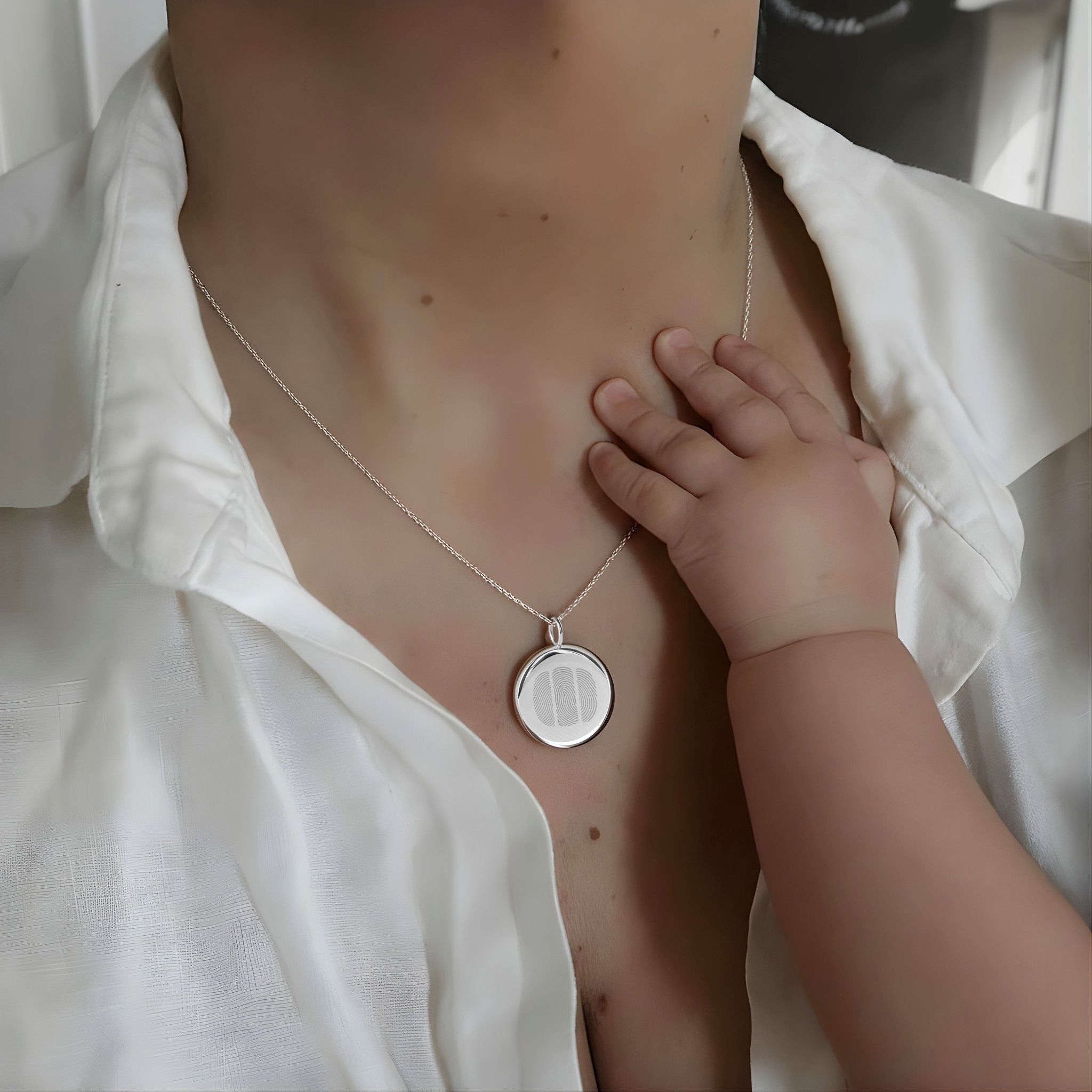 Emotional portrait of a mother wearing the Danibydsgn Three Fingerprint Coin Necklace in 925 Sterling Silver as her baby’s hand rests gently on her chest — symbolising family, love, and everlasting connection captured in timeless craftsmanship.