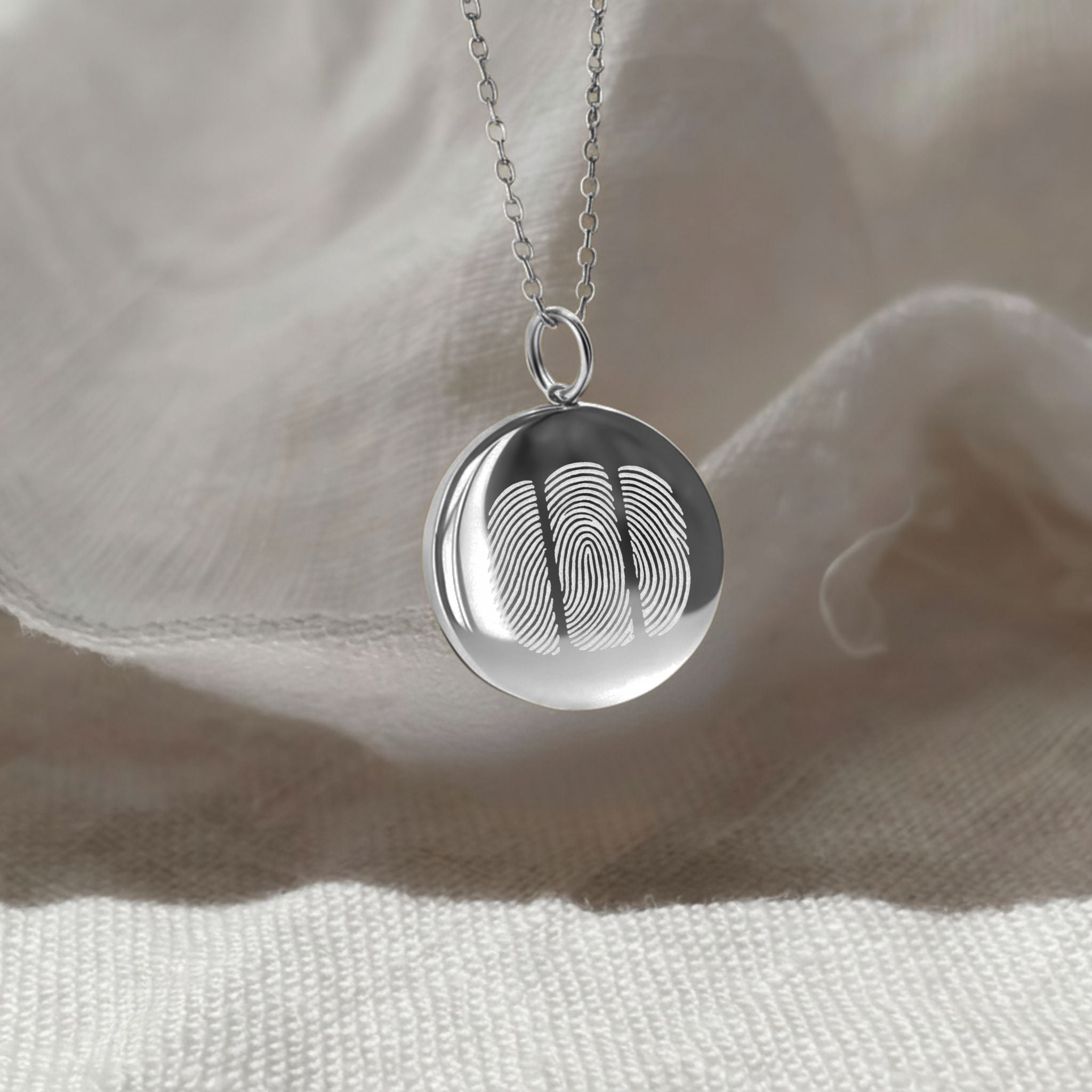 The Danibydsgn Three Fingerprint Coin Necklace in 925 Sterling Silver resting gently against soft fabric, illuminated by natural light — symbolising family, love, and unity through three beautifully engraved fingerprints.