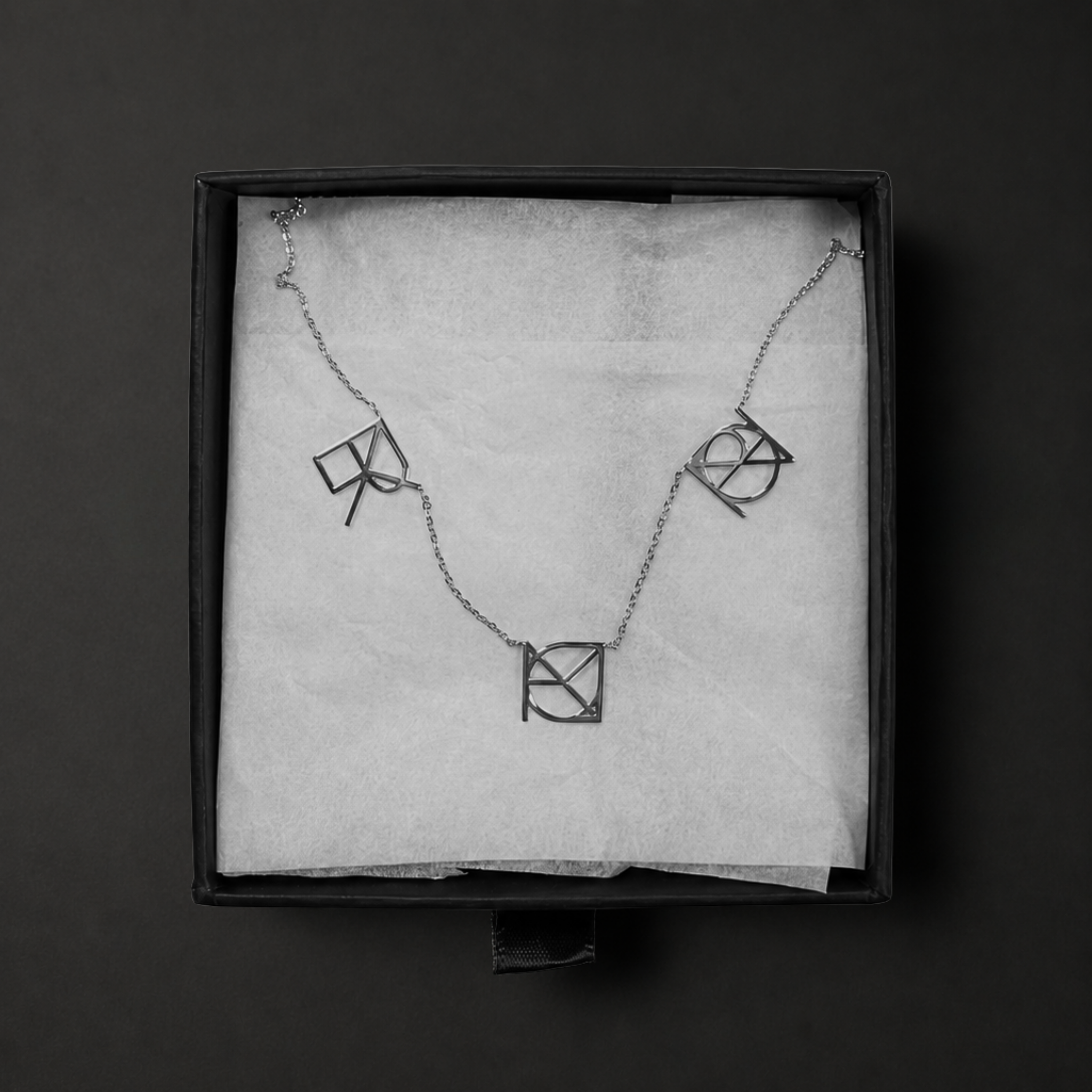 A top-down product shot of the Danibydsgn three symbol pendant necklace in 925 sterling silver presented flat inside a black jewelry gift box. This personalized meaningful jewelry piece features three cut-out uppercase geometric charms handcrafted from a custom fineline Danibydsgn design, arranged elegantly on textured white tissue paper.