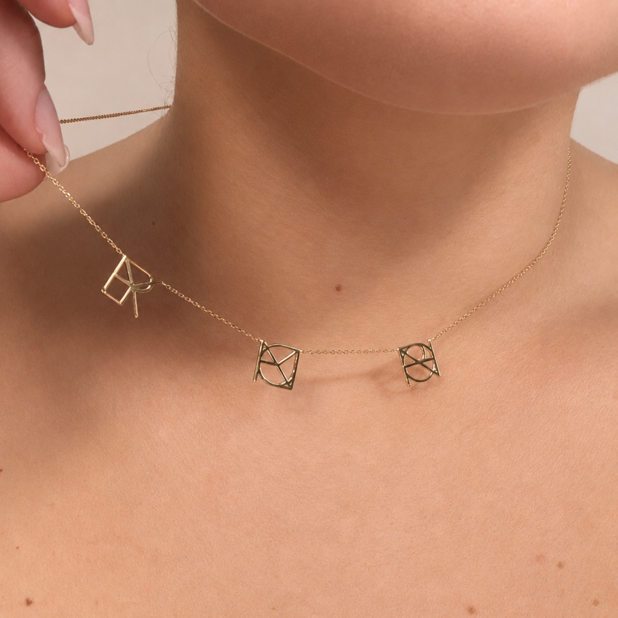A close-up lifestyle image of a model's neck as her fingers gently lift the chain of a Danibydsgn three symbol pendant necklace in 9k solid gold. This made to order modern heirloom features three personalized uppercase geometric charms handcrafted from unique names, words, or dates on a delicate yellow gold chain.