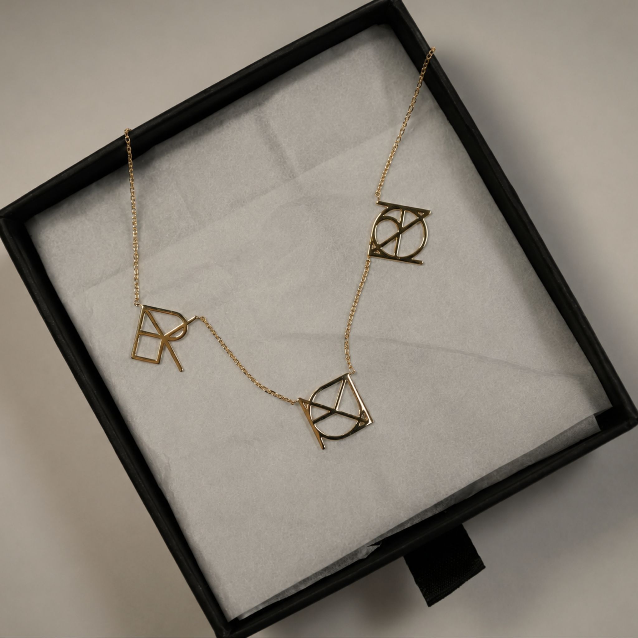 The Danibydsgn three symbol pendant necklace in 9k solid gold beautifully presented inside a premium black gift box. This made to order modern heirloom necklace features three personalized uppercase geometric charms handcrafted from meaningful names, words, or dates, resting flat on white tissue paper to showcase the unique custom fine jewelry design.