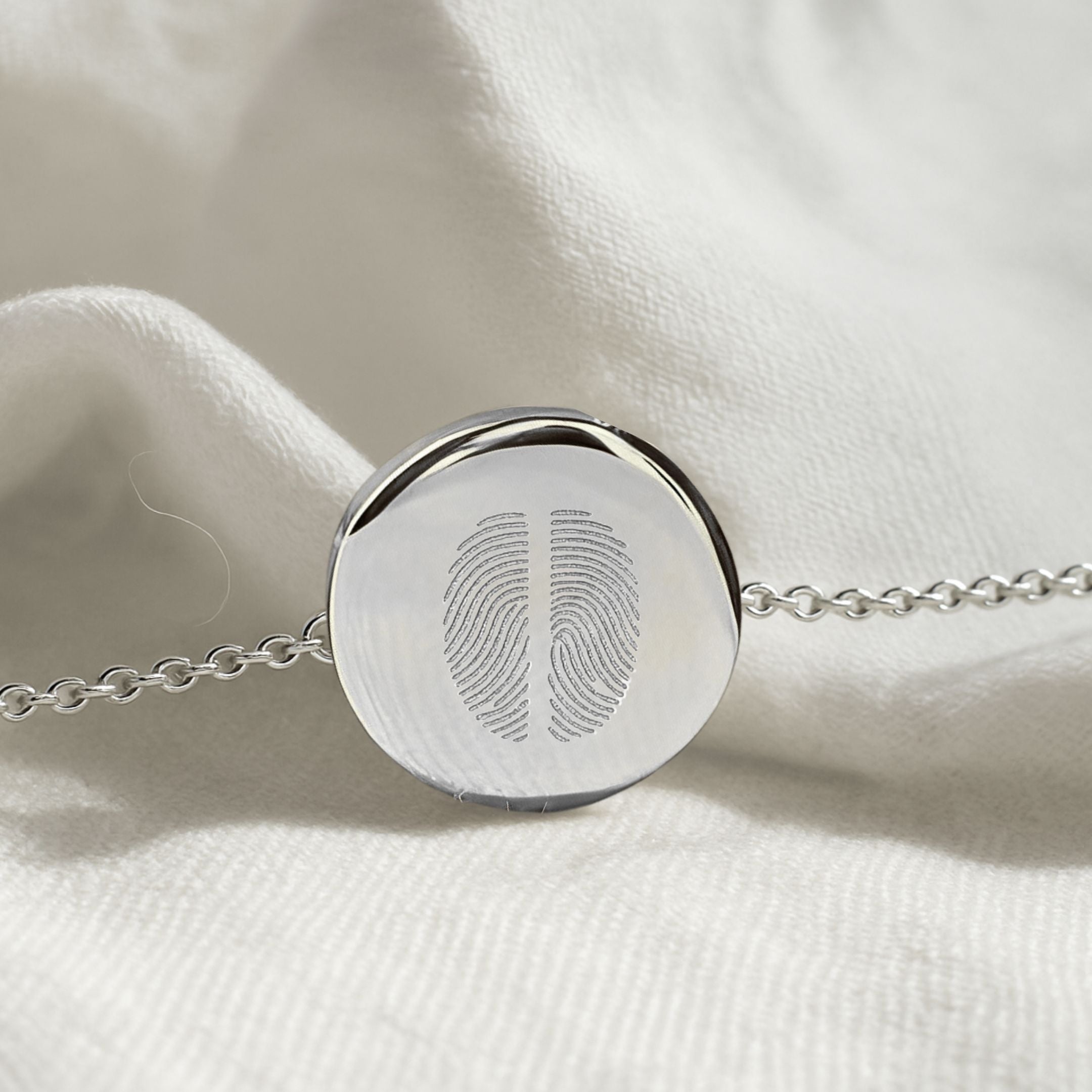 Close-up of the Danibydsgn Two Fingerprint Coin Bracelet in 925 Sterling Silver resting on soft white fabric, highlighting the dual fingerprint engraving and polished coin pendant detail.