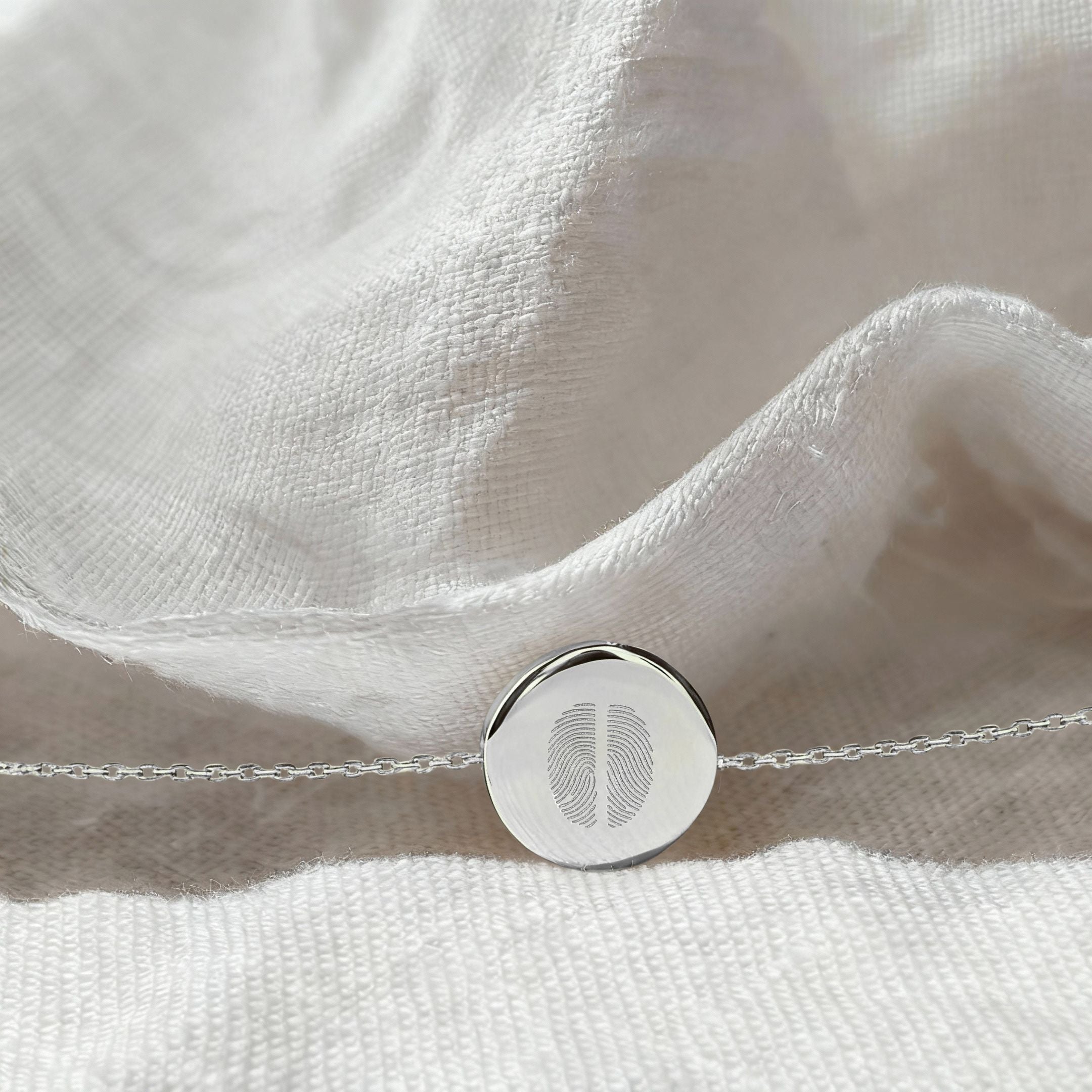 The Danibydsgn Two Fingerprint Coin Bracelet in 925 Sterling Silver resting on softly textured white fabric, highlighting the detailed dual fingerprint engraving and polished round pendant.