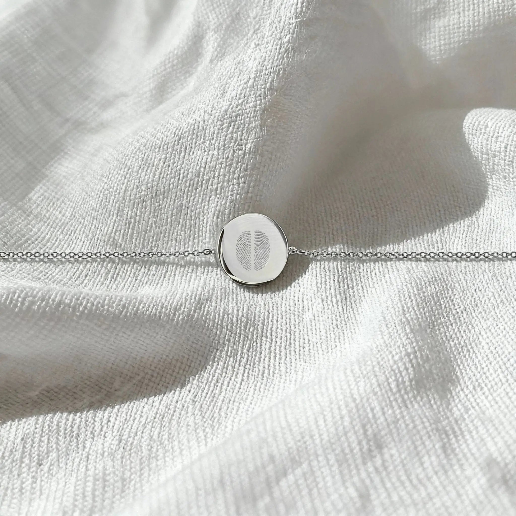 The Danibydsgn Two Fingerprint Coin Bracelet in 925 Sterling Silver displayed flat on soft white fabric, showcasing the delicate dual fingerprint engraving and elegant minimalist chain.