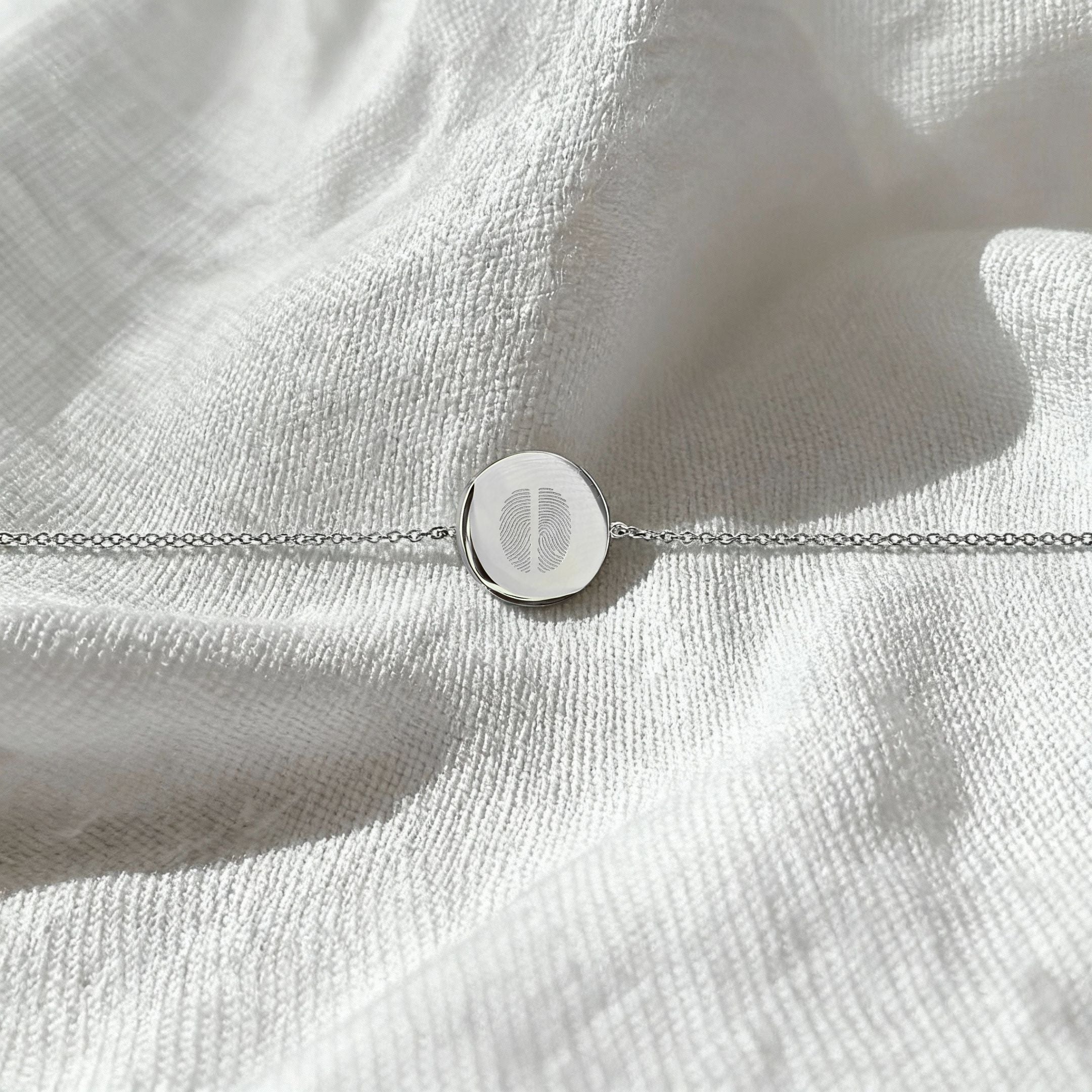 The Danibydsgn Two Fingerprint Coin Bracelet in 925 Sterling Silver displayed flat on soft white fabric, showcasing the delicate dual fingerprint engraving and elegant minimalist chain.