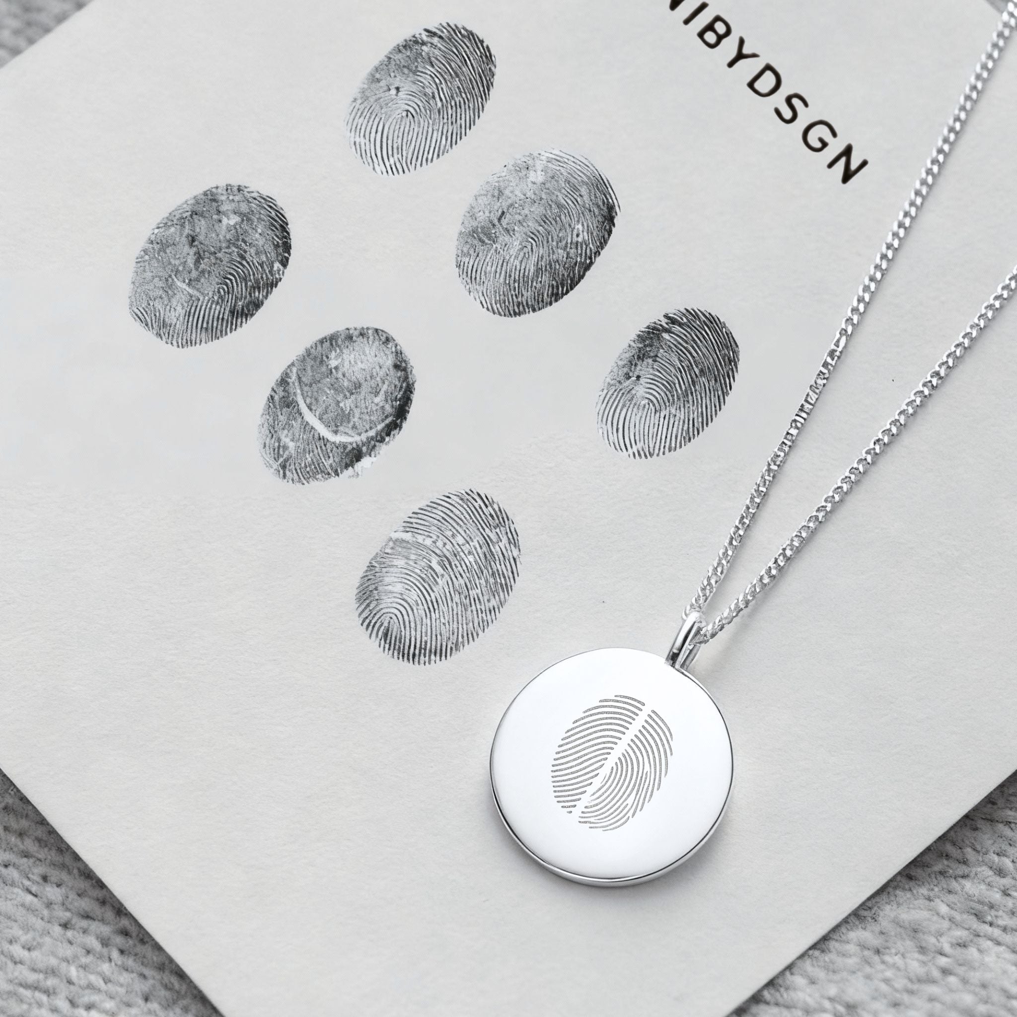 The Danibydsgn Two Fingerprint Coin Necklace in 925 Sterling Silver placed beside real fingerprint imprints — a beautiful reflection of how each piece is thoughtfully engraved to preserve the unique bond and touch between two loved ones.