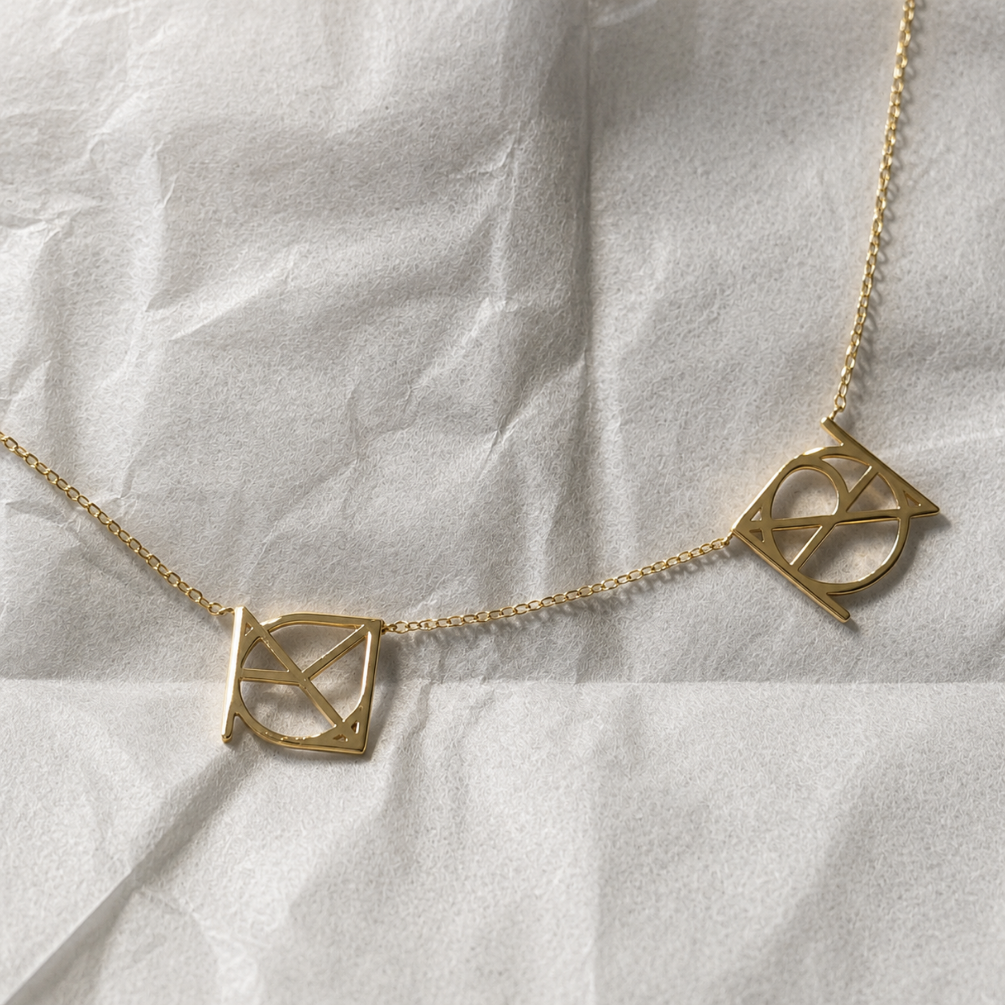 A top-down studio product shot of the Danibydsgn two symbol pendant necklace in 9k solid gold. This made to order modern heirloom necklace features two personalized uppercase geometric charms handcrafted from a customer's meaningful names, words, or dates, elegantly displayed across a textured white crinkled paper background.
