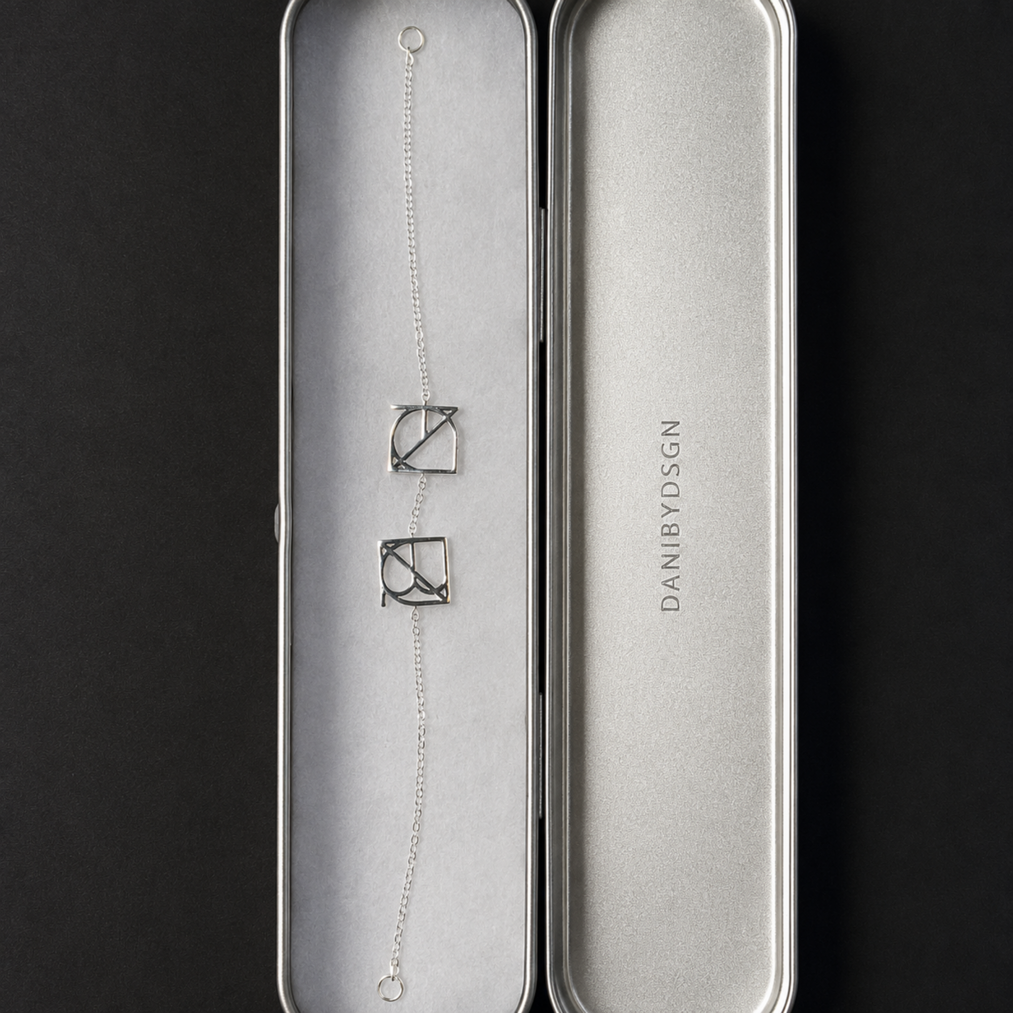 A top-down product shot of the Danibydsgn two symbol pendant bracelet in 925 sterling silver displayed vertically inside its open, brushed silver metal gift case. This personalized meaningful jewelry piece showcases the two custom uppercase geometric charms resting on protective white padding alongside the vertically embossed brand name lid on a black background.