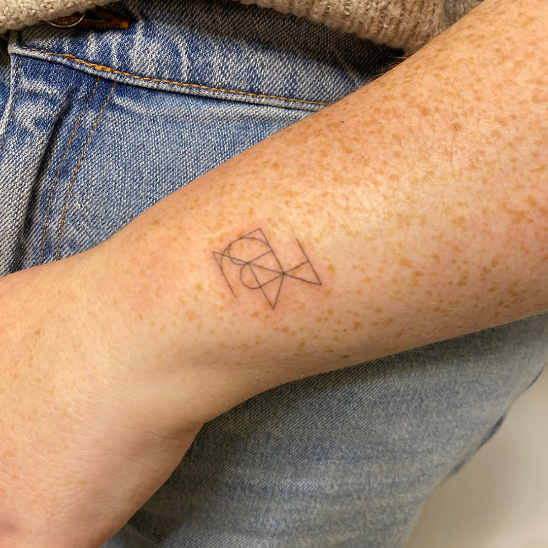 Joined custom logo and fine line tattoo design showing two geometric symbols combined into one minimal artwork on a lady's wrist.
