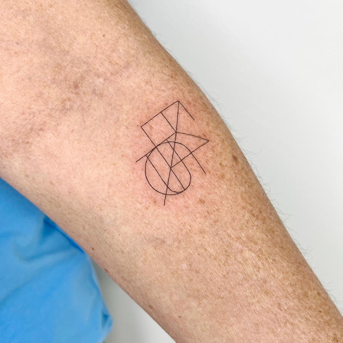 Joined fine line tattoo design featuring two geometric symbols combined into a custom logo artwork on forearm.