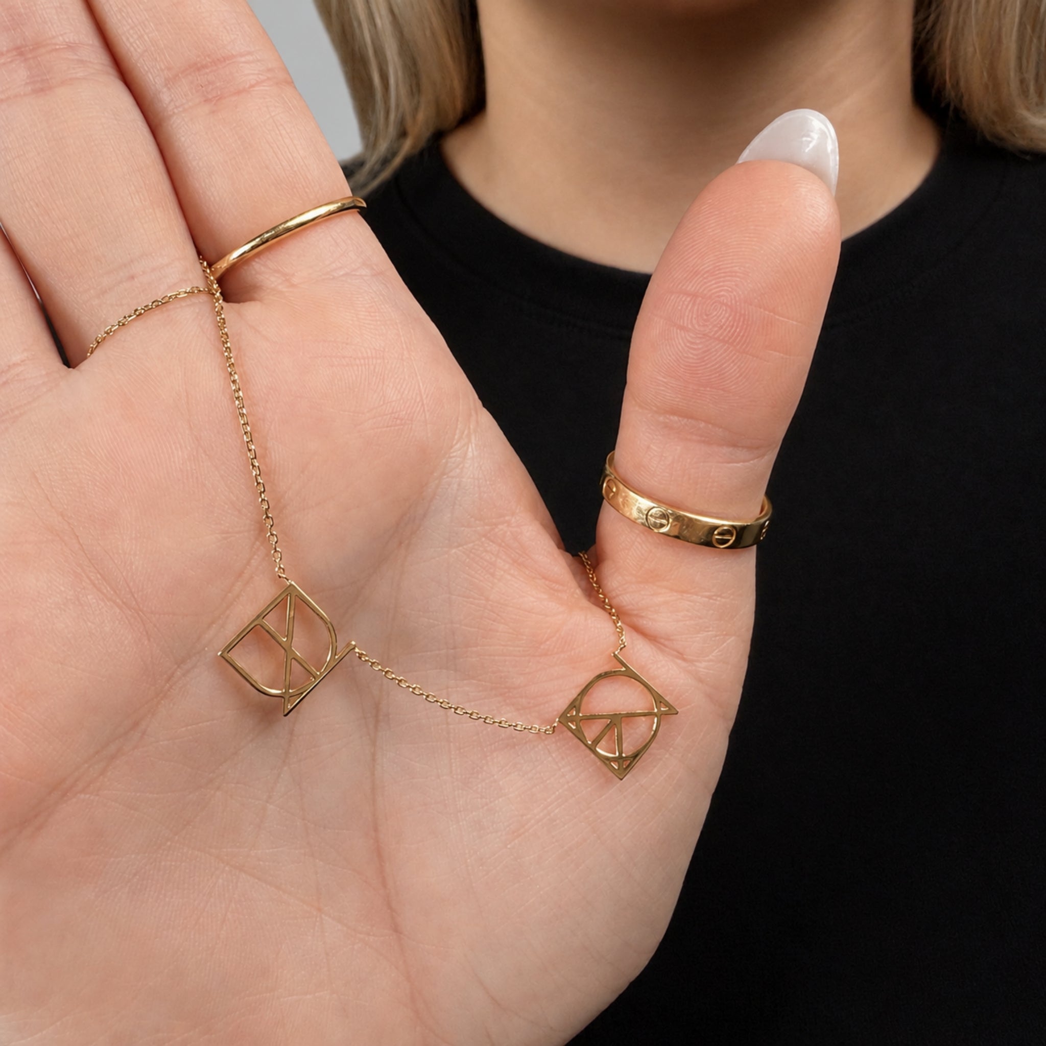 Close-up detail of a custom Danibydsgn two symbol necklace in solid 9k gold held in a hand, showcasing two unique cut out jewelry symbol hand-designed from personal stories on a fine chain.