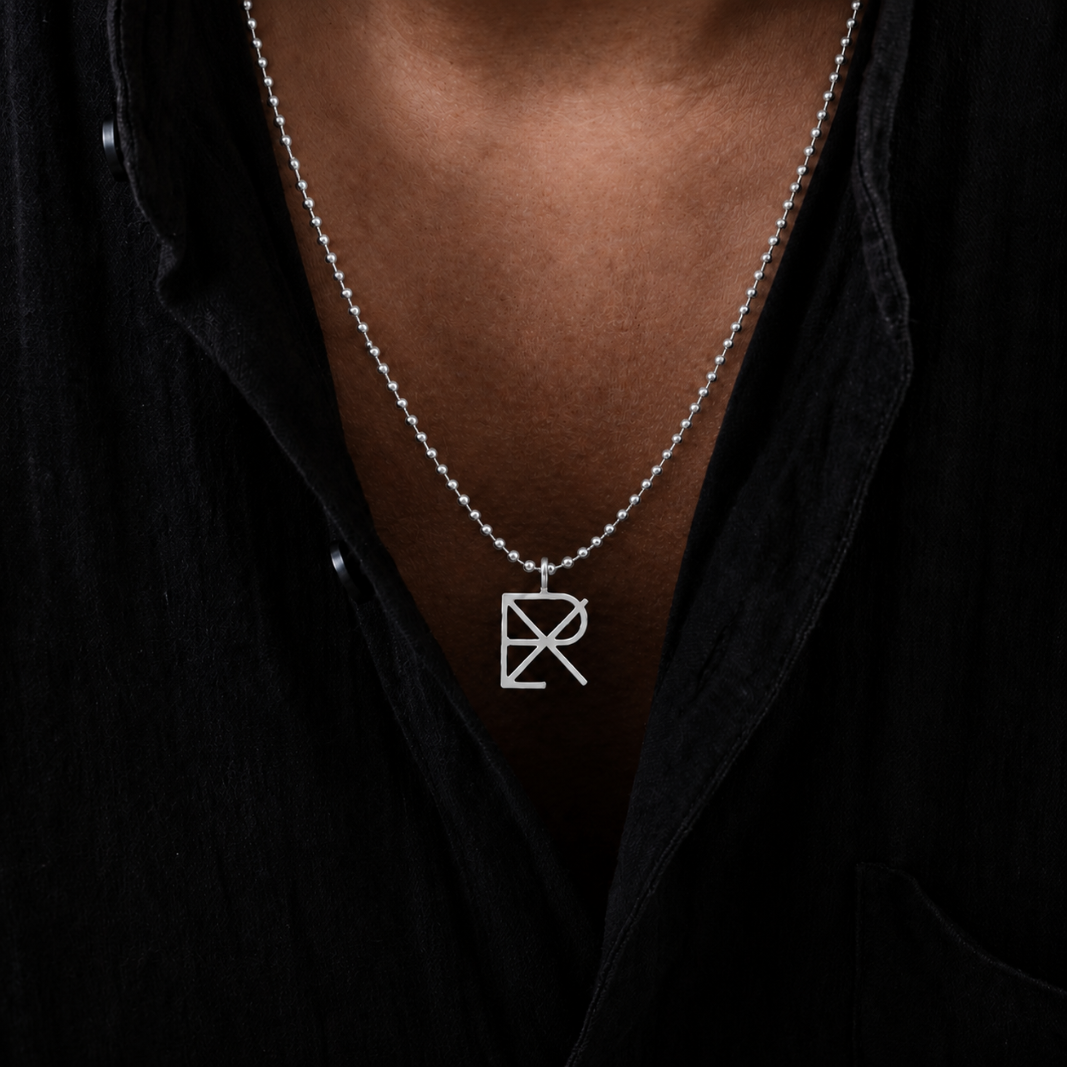 A gold ball chain necklace with a Danibydsgn custom cut out symbol pendant necklace in 925 sterling silver, worn by a man in a black t-shirt.  Longer chain that sits chest level.