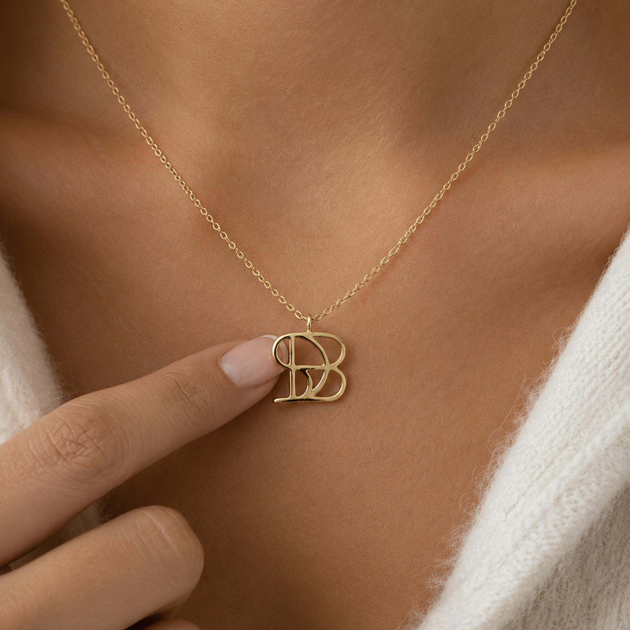 A close-up lifestyle image of a model wearing the Danibydsgn single symbol pendant necklace in 9k solid gold. The model's finger gently touches a personalized uppercase geometric initial charm, handcrafted from a custom line art design, hanging on a delicate yellow gold chain over the collarbone while styled with a soft white knit sweater.