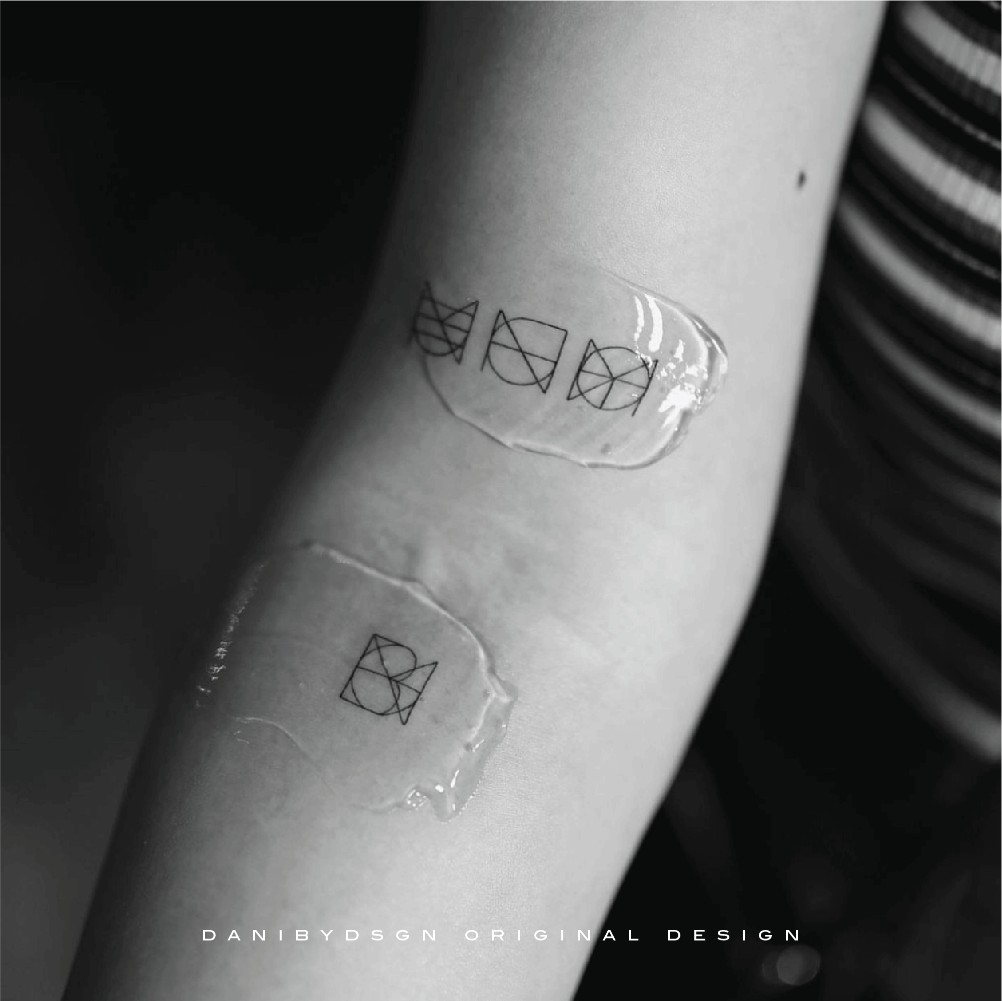 A monochromatic image displaying a fineline tattoo by Danibydsgn, with three intricate symbols accompanied by a single emblem, freshly inked on the forearm and sealed with protective wrapping. This minimalist name logo design is a modern aesthetic as personal branding, a hallmark of Danibydsgn's custom name logo tattoo designs.  The geometric tattoo designs are both minimalist and meaningful yet elegant and discrete.  Danibydsgn is the OG of personal name logo designs