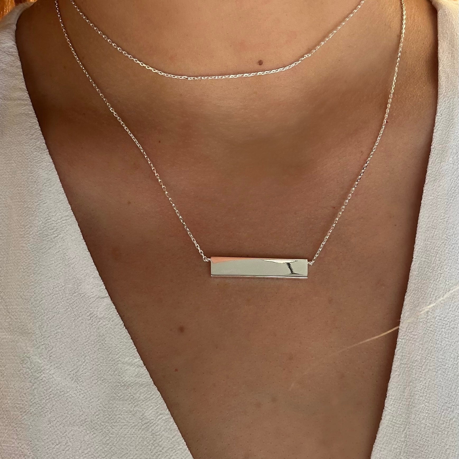 This photo is of a sleek Engravable Silver Bar Necklace which is around the neck of a lady and elegantly rests just below the collarbone.  The high-polish finish captures light beautifully, making it a versatile piece for both personal branding and meaningful gifts. This necklace is made for customized engraving with a Danibydsgn's custom logo design turning a simple silver bar necklace into a unique piece of personalized jewelry, perfect as a keepsake jewelry item or a personalized gift. 